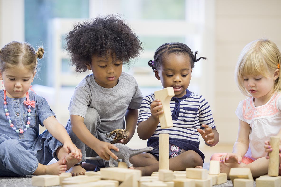 A multi-ethnic group of toddlers are sitting together on the floor holding playing with wood blocks together.